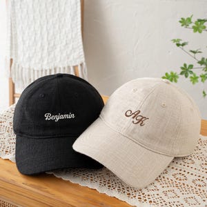 May include: A black baseball cap with the name Benjamin embroidered in white thread and a beige baseball cap with the initials AK embroidered in brown thread. Both caps feature a textured fabric finish and a structured six-panel design.