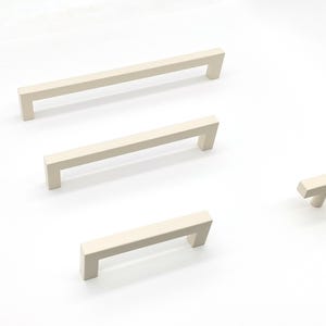 Op de afbeelding: Een set van vier matte gebroken witte kastgrepen en één T-vormige knop. De set bevat drie rechthoekige stanggrepen van verschillende lengtes en één kleine T-knop, allemaal met een gladde afwerking en strakke, rechte randen.