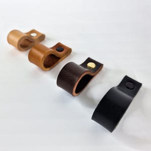 May include: Four leather drawer pulls in varying shades of brown and black. Each pull is a loop design with a metal rivet. The colors range from light tan to dark brown and black.