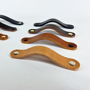 May include: A collection of leather drawer pulls in various shades of brown and black. Each pull has a curved design with rounded ends and is secured with metal rivets. The hardware is suitable for cabinets, drawers, or other furniture.