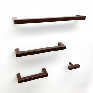 May include: A set of four dark brown cabinet hardware pieces, including three rectangular bar pulls of varying lengths and one T-shaped knob, all featuring a smooth, matte finish and clean, angular lines for furniture or cabinetry applications.