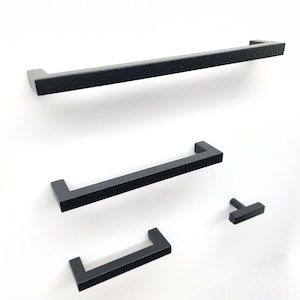 May include: A collection of four matte black cabinet hardware pieces, including three rectangular bar pulls of varying lengths and one T-shaped knob. Each piece features a clean, linear design with sharp edges and a consistent dark finish.