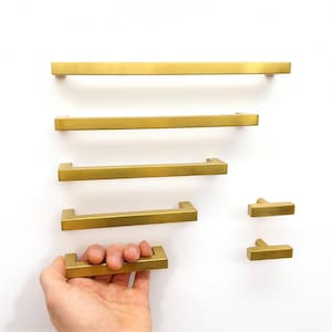 Può includere: Una collezione di sei maniglie e pomelli per armadietti in oro spazzolato di varie lunghezze. La ferramenta presenta un design lineare e rettangolare con una finitura metallica liscia e opaca. Gli articoli sono disposti in colonna e in fila.
