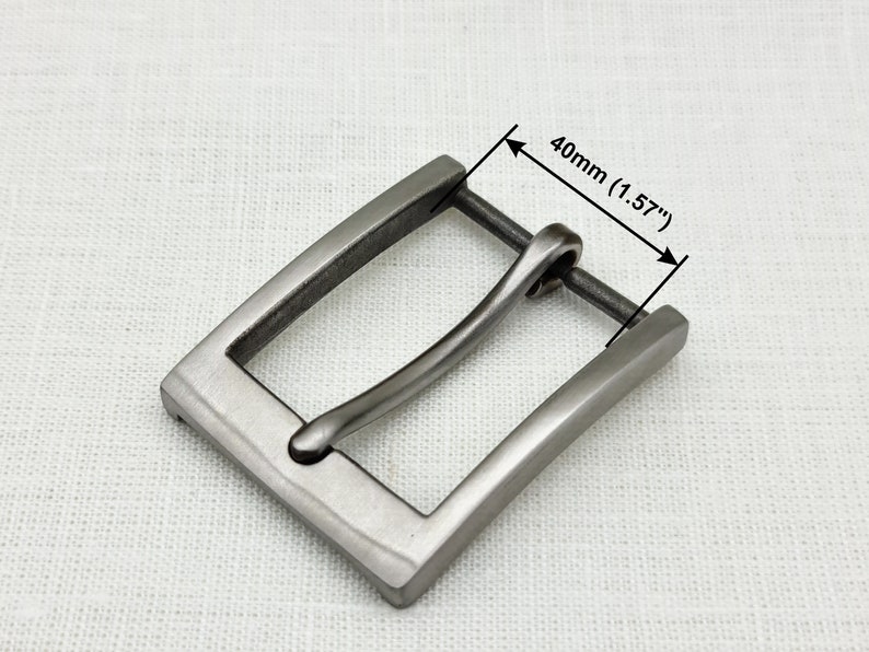May include: A silver-toned metal belt buckle with a rectangular frame and a curved, smooth buckle tongue. The buckle measures 1.57 inches (40 mm) wide.