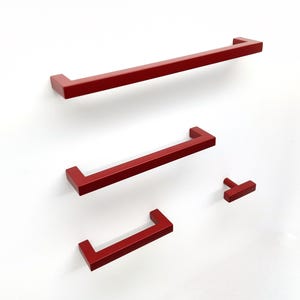 May include: A collection of four matte red cabinet hardware pieces, including three rectangular bar pulls of varying lengths and one small T-shaped knob, all featuring a clean, angular design with flat surfaces and sharp, defined edges.