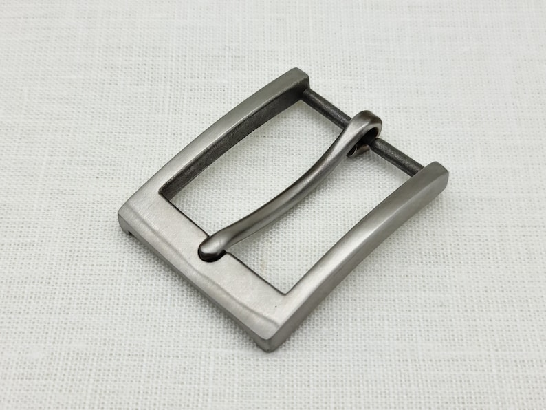 May include: A silver-toned metal belt buckle with a rectangular frame and a curved, smooth pin.