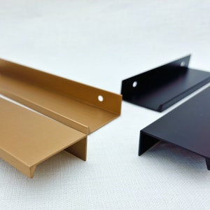 Puede incluir: Una selección de tiradores de armario modernos en varios colores. Los tiradores son rectangulares con un labio y un agujero para el montaje. Los colores incluyen dorado, negro y bronce oscuro. Los tiradores son de metal.