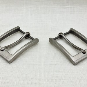 May include: Two silver-toned metal belt buckles with a curved, smooth, and slightly rounded design. The buckles are rectangular with a slightly curved edge.