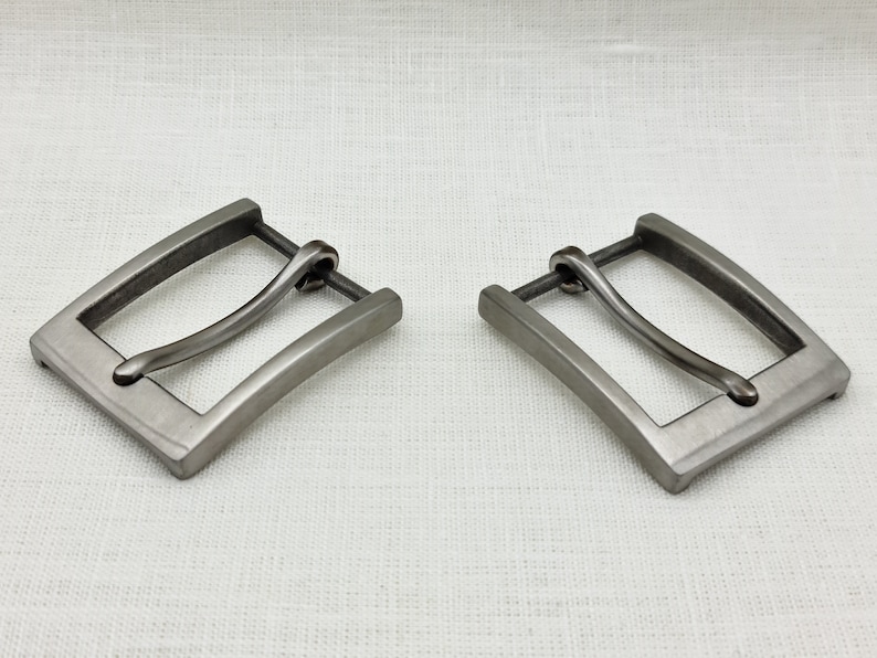 May include: Two silver-toned metal belt buckles with a curved, smooth, and slightly rounded design. The buckles are rectangular with a slightly curved edge.