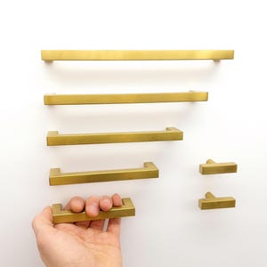 Peut inclure: Une collection de poignées de placard en or brossé comprenant quatre barres rectangulaires de longueurs variées et deux petits boutons en forme de T. Les poignées métalliques présentent un design linéaire plat avec une finition lisse.