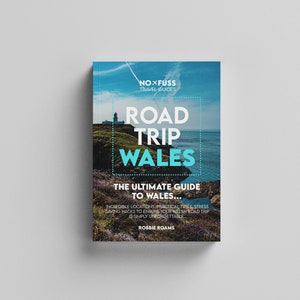 May include: A book cover with a blue and white design featuring a lighthouse and ocean. The title is "Road Trip Wales: The Ultimate Guide to Wales..." with the subtitle "Incredible locations, practical tips & stress saving hacks to ensure your Welsh road trip is simply unforgettable." The author is Robbie Roams.