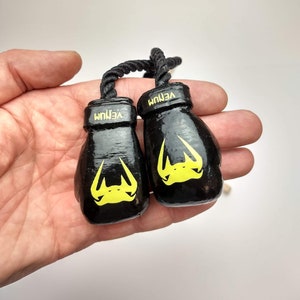 Puede incluir: Un par de guantes de boxeo negros con la marca Venum en amarillo. Los guantes est&aacute;n atados con una cuerda negra.