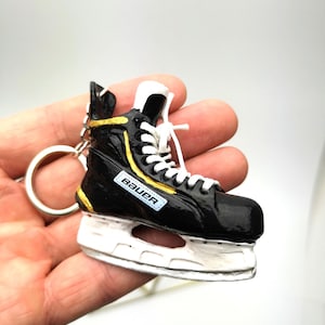 Op de afbeelding: Miniatuur zwarte en witte ijsschaatssleutelhanger met gouden accenten. De sleutelhanger heeft witte veters en de merknaam "Bauer". Het mes is wit en de schaats is bevestigd aan een zilveren sleutelring.