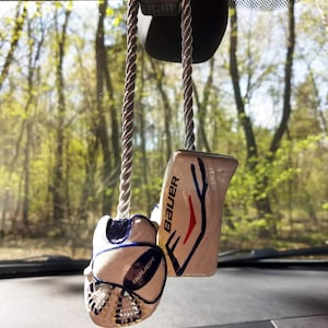 May include: A white and blue hockey glove and a white hockey stick with red and blue accents hanging from a rearview mirror in a car. The hockey stick has the word "Bauer" written on it.