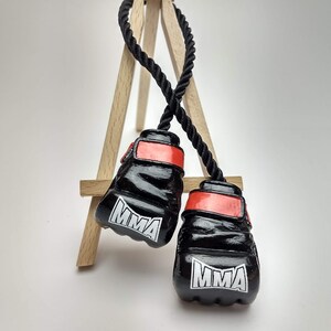 以下が含まれることがあります： 赤い縁取りと白い文字で「MMA」と書かれた黒いボクシンググローブのペア。グローブは木製のイーゼルに黒いロープで吊るされています。