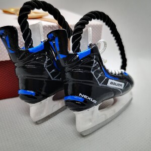 May include: A pair of black and blue hockey skate ornaments with white blades. The skates are decorated with a white and gray pattern and the words "Bauer" and "Nexus".
