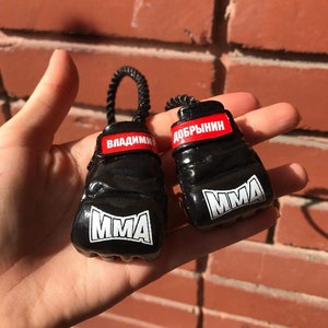 以下が含まれることがあります： 赤と白の文字が入った黒いボクシンググローブのキーホルダー。グローブの文字は「MMA」と「VLADIMIR DOBRYNIN」です。
