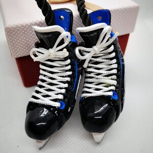 May include: A pair of black and blue miniature ice skates with white laces. The skates have the word "Bauer" printed on them.