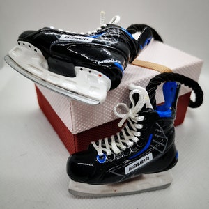 May include: Two black and white hockey skate ornaments with blue accents. The skates are tied with white laces and have the word "Bauer" printed on them.