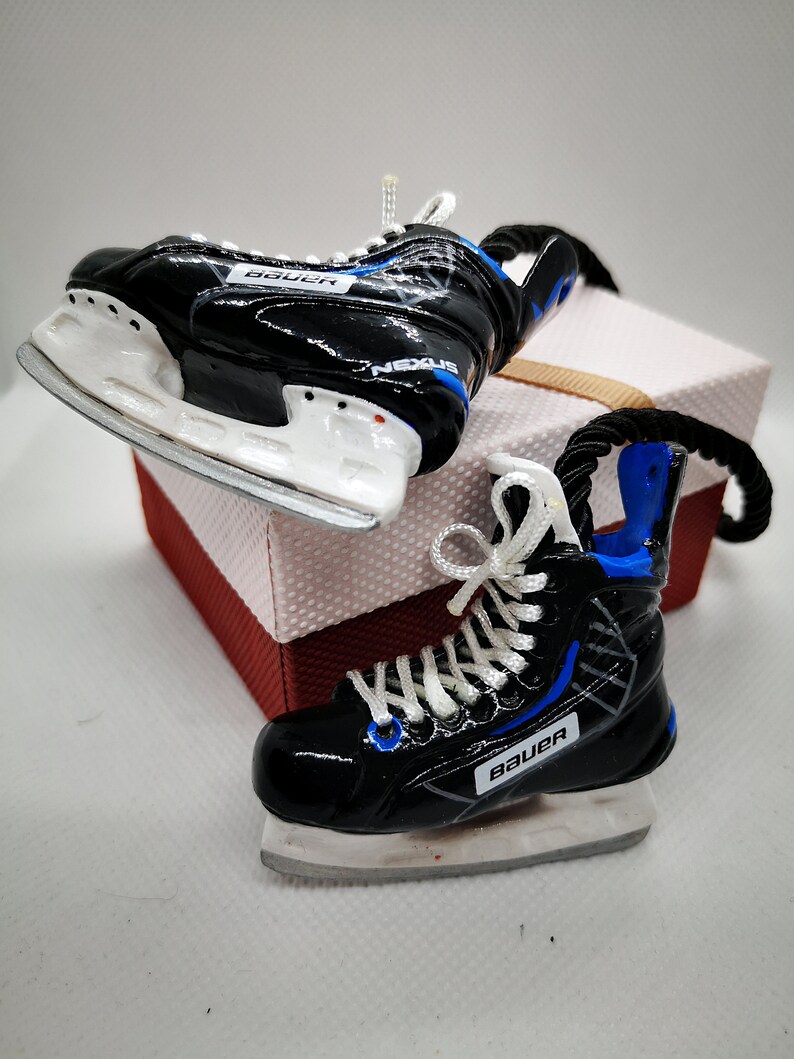 May include: Two black and white hockey skate ornaments with blue accents. The skates are tied with white laces and have the word "Bauer" printed on them.