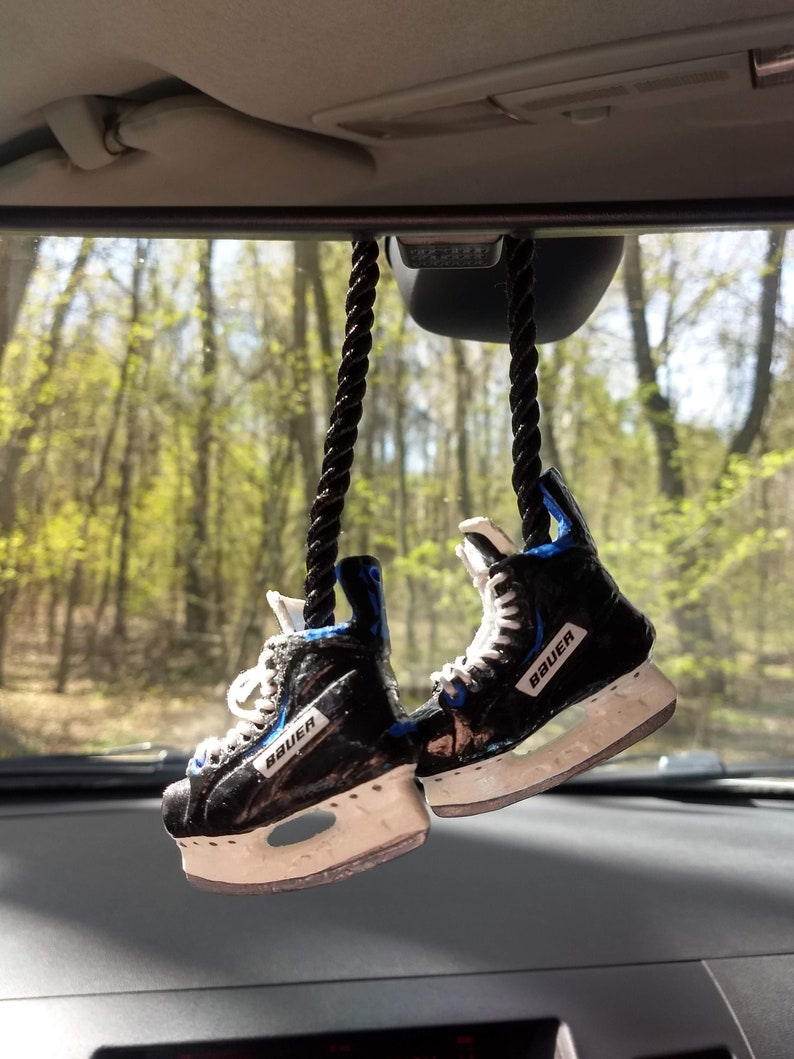 May include: A pair of black and white ice skates with blue accents hanging from a rearview mirror in a car. The skates have the word "Bauer" printed on them.