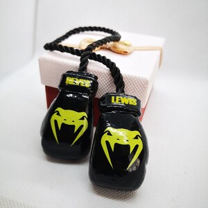 Puede incluir: Llaveros de guantes de boxeo negros con las palabras "REYES" y "LEWIS" en ellos. Los guantes tienen un logotipo de serpiente amarilla.
