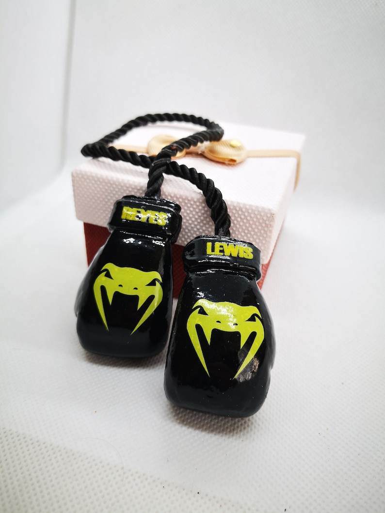 Puede incluir: Llaveros de guantes de boxeo negros con las palabras "REYES" y "LEWIS" en ellos. Los guantes tienen un logotipo de serpiente amarilla.