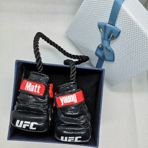 May include: A pair of black boxing gloves with red trim and white lettering. The gloves are in a blue box with a black rope handle. The gloves have the names "Matt" and "Young" on them, as well as the letters "UFC".