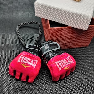 May include: A pair of pink and black Everlast boxing gloves with a black cord attached. The gloves are on a black surface with a red and white gift box in the background.