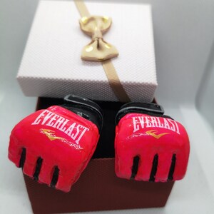 May include: A pair of pink boxing gloves with black trim and the word "EVERLAST" in white lettering. The gloves are in a brown gift box with a white lid and a beige bow.