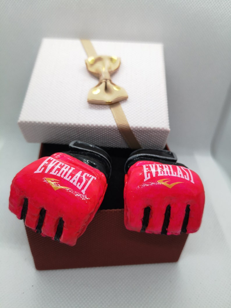May include: A pair of pink boxing gloves with black trim and the word "EVERLAST" in white lettering. The gloves are in a brown gift box with a white lid and a beige bow.
