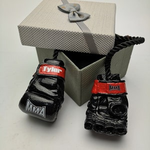 May include: A pair of black boxing gloves with red trim and the word "MMA" printed on them. The gloves are attached to a black rope and are in a gray gift box with a gray bow.