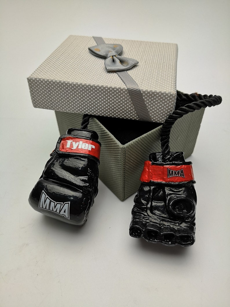 May include: A pair of black boxing gloves with red trim and the word "MMA" printed on them. The gloves are attached to a black rope and are in a gray gift box with a gray bow.