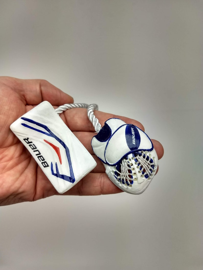 May include: A white and blue hockey goalie set with a white and blue hockey stick charm. The stick charm has the word "Bauer" printed on it. The goalie set has a white glove with a blue Bauer logo and a white blocker with a blue Bauer logo.