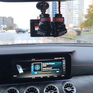 May include: A black car interior with a rearview mirror. Hanging from the mirror are two black boxing gloves with red accents. The gloves have the names "Conor" and "Khabib" written on them. The gloves are attached to red strings and have the word "UFC" written on them.