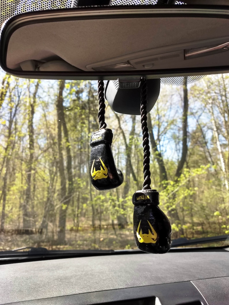 Puede incluir: Dos ambientadores de coche en forma de guantes de boxeo negros colgados del espejo retrovisor de un coche. Los guantes tienen detalles amarillos y la palabra "VENUM" est&aacute; impresa en uno de los guantes.