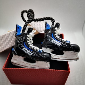 May include: A pair of black and blue hockey skate ornaments with white blades and a black and white Bauer logo. The ornaments are hanging from a black rope and are in a red box.