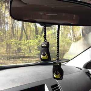 Puede incluir: Dos ambientadores de coche en forma de guantes de boxeo negros colgados del espejo retrovisor de un coche. Los guantes tienen detalles amarillos y la palabra "VENUM" est&aacute; impresa en ellos.