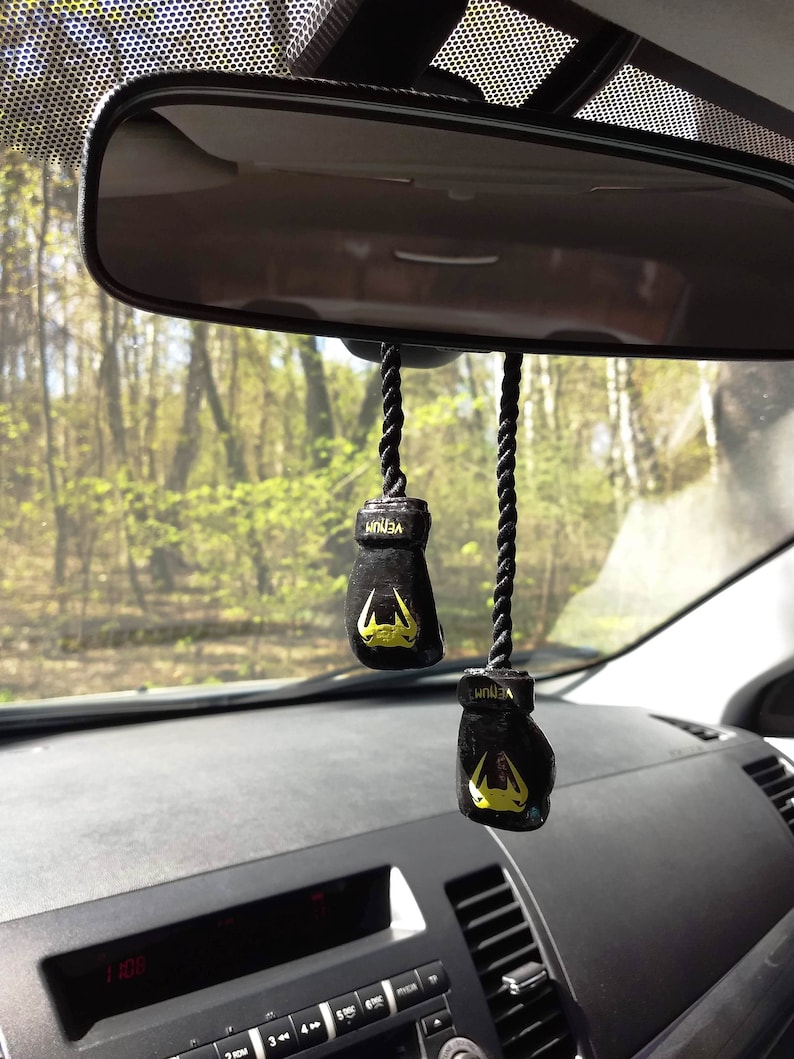 Puede incluir: Dos ambientadores de coche en forma de guantes de boxeo negros colgados del espejo retrovisor de un coche. Los guantes tienen detalles amarillos y la palabra "VENUM" est&aacute; impresa en ellos.