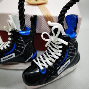 May include: A pair of black and blue ice hockey skate ornaments with white laces and the word "Bauer" on each skate. The skates are hanging from a black rope.