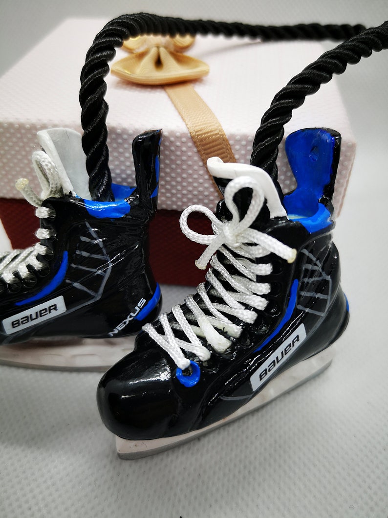 May include: A pair of black and blue ice hockey skate ornaments with white laces and the word "Bauer" on each skate. The skates are hanging from a black rope.