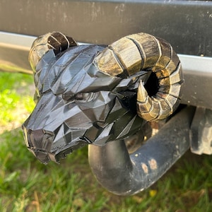 May include: A black, low-poly ram head hitch cover featuring large, curved horns with a distressed gold-tone finish. The geometric, faceted design of the ram head is mounted onto a vehicle trailer hitch receiver, showcasing a sharp, angular aesthetic.