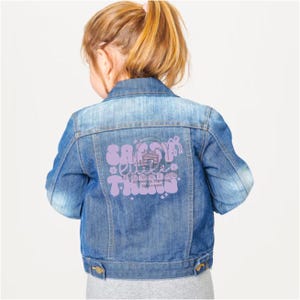 May include: A light blue denim jacket with a purple graphic on the back that says "Sassy Little Things". The graphic includes a cabin illustration and the words "Embroidery, Engravings, & more".