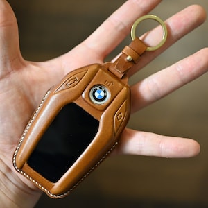 May include: A brown leather key fob with a silver key ring and a blue BMW logo. The fob has a stitched edge and a small screen.