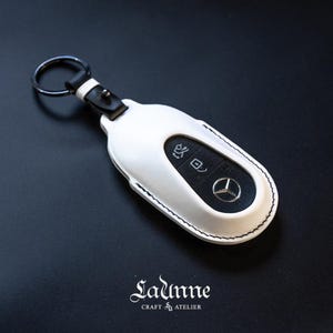 May include: White leather key fob with a black Mercedes-Benz logo and buttons. The fob has a black leather strap with a silver ring.