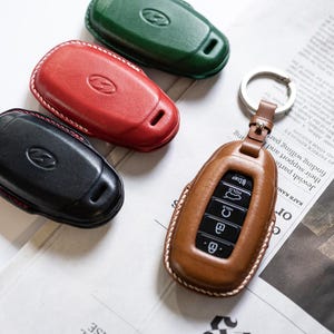 Pode incluir: Capas de chaveiro de couro em várias cores: castanho, vermelho, preto e verde. Cada capa tem uma borda costurada e um logotipo de carro. Uma capa castanha está presa a um porta-chaves. As capas de chaveiro estão em um jornal.