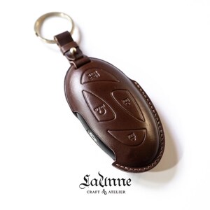 May include: A brown leather key fob with a key ring. The fob has four buttons with raised lettering. The text on the fob reads "Ladunne Craft & Atelier".