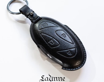 May include: Black leather key fob with a key ring and four buttons. The fob is designed to protect a car key.