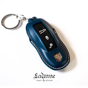 Puede incluir: Un llavero de cuero azul con costuras blancas y un logotipo de Porsche. El llavero tiene un anillo de llave plateado y cuatro botones.