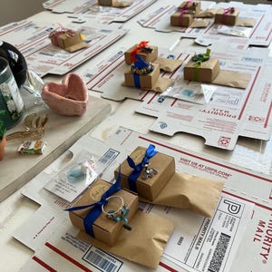 May include: A collection of small, wrapped packages containing jewelry. The packages are arranged on a white surface with brown cardboard boxes and a heart-shaped dish. The packages are tied with blue, green, and red ribbon. The packages are labeled with "PRIORITY MAIL" and "VISIT US AT USPS.COM ORDER FREE SUPPLIES ONLINE".